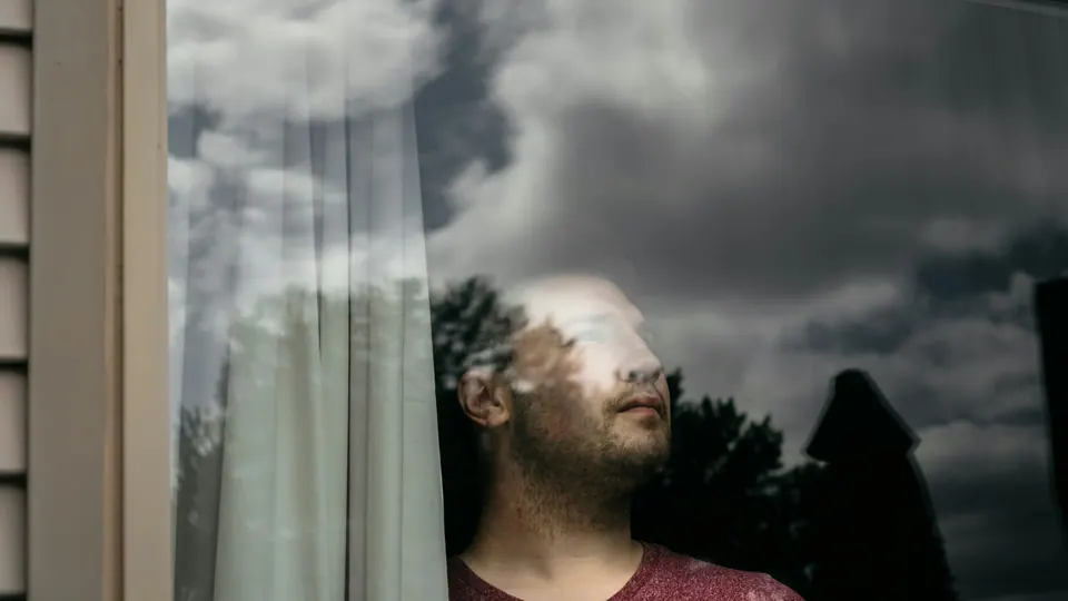 Homem olhando pela janela num dia nublado, com o céu refletido no vidro